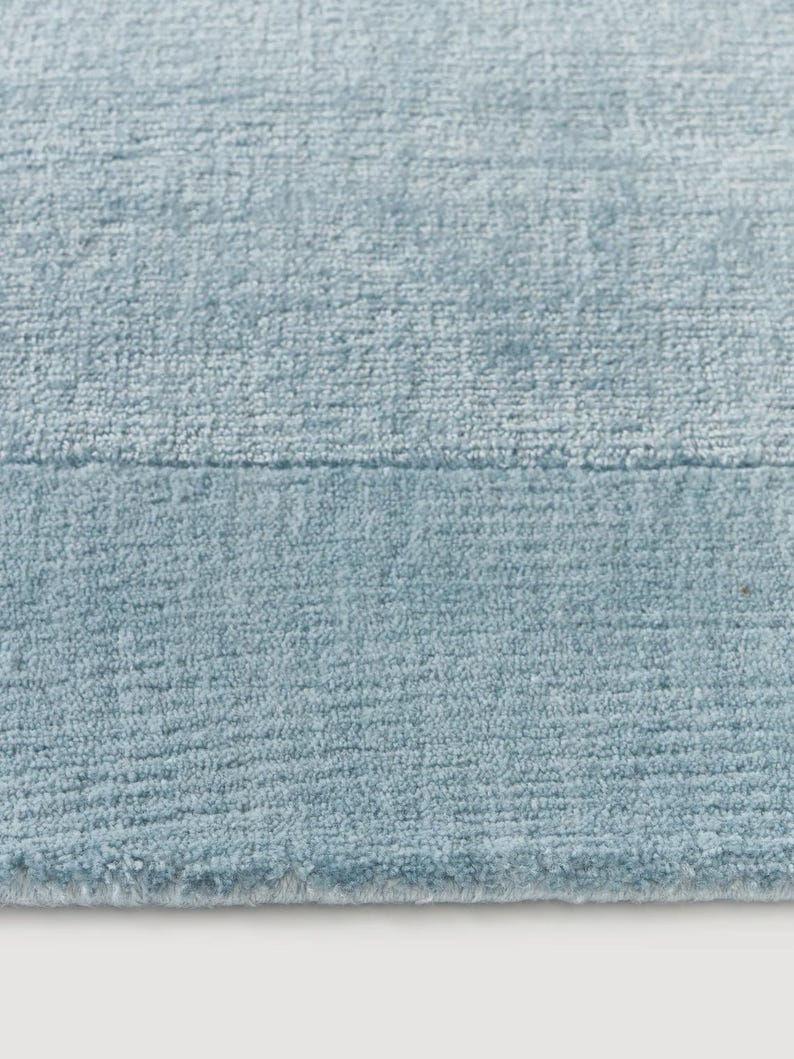 May include: Close-up of a light blue rug with a textured surface. The rug has a soft, plush appearance and a subtle color variation. The texture is visible, with a slight sheen.