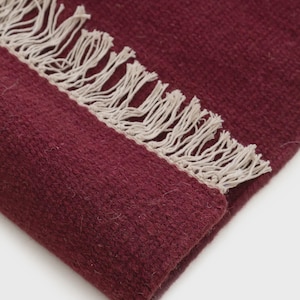 May include: A deep burgundy woven textile featuring a thick, textured surface and a prominent row of off-white fringe along the edge. The fabric displays a dense, consistent weave pattern throughout, with the light-colored tassels creating a contrast.