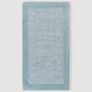 May include: A rectangular, light blue rug with a textured surface. The rug features a lighter blue rectangular border that frames the central area. The rug is designed for home decor.