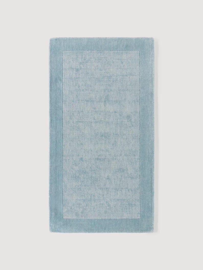 May include: A rectangular, light blue rug with a textured surface. The rug features a lighter blue rectangular border that frames the central area. The rug is designed for home decor.