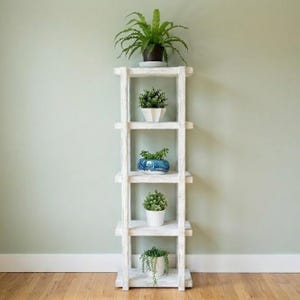 Puede incluir: Un estante blanco de madera para plantas con cuatro estantes, cada uno con una planta en maceta. Las plantas varían en tamaño y tipo, con follaje verde. El estante está contra una pared verde claro y se encuentra sobre un piso de madera.