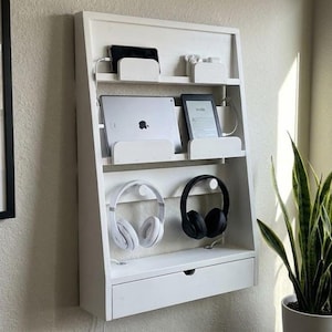 May include: White wooden wall-mounted organizer with multiple shelves and a drawer. It holds a tablet, e-reader, phone, and headphones. The organizer has a clean, modern design, perfect for a home office or workspace. The headphones are white and black.