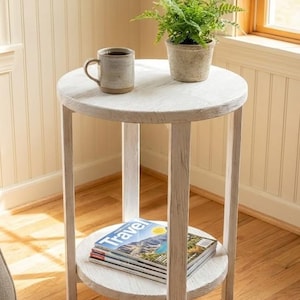 May include: A white, round side table with a lower shelf, holding a stack of magazines. A mug and a potted fern sit on the top surface. The table has a distressed, rustic finish, and is placed in a room with wood flooring.