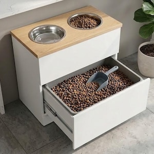 May include: A white pet food station with a light wood top. It has two stainless steel bowls and a drawer filled with pet food. A gray scoop rests in the drawer. A potted plant is in the background.