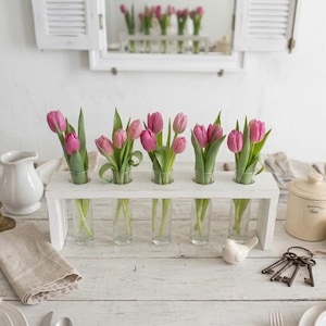 May include: A white wooden flower holder with five glass vases, each holding pink tulips. The table setting includes brass candlesticks, a white pitcher, a ceramic jar, and a set of keys.