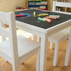 May include: A white wooden children's table with a black chalkboard top, surrounded by two matching chairs. Colorful chalk sticks and an eraser are scattered on the table's surface, suggesting creative play. The table is designed for young children.