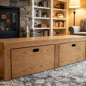 May include: A rectangular, light-brown wooden coffee table with two drawers. The drawers have recessed handles. The table is placed on a patterned rug in front of a fireplace and a bookshelf.