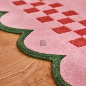 May include: A close-up of a pink rug with a scalloped edge and a red checkered pattern. The edge is trimmed with green. The rug is on a wooden floor.