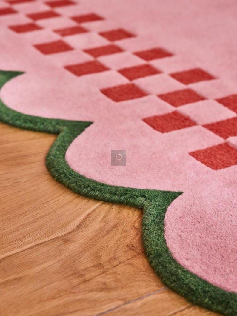 May include: A close-up of a pink rug with a scalloped edge and a red checkered pattern. The edge is trimmed with green. The rug is on a wooden floor.