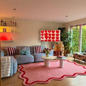 May include: A living room with a light blue sofa, a pink rug with a red border, and a white coffee table. A TV displays red hearts. Decorative pillows and plants add to the room's aesthetic.