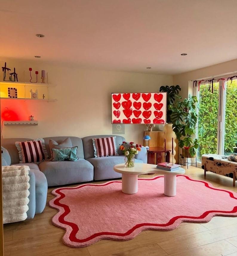 May include: A living room with a light blue sofa, a pink rug with a red border, and a white coffee table. A TV displays red hearts. Decorative pillows and plants add to the room's aesthetic.