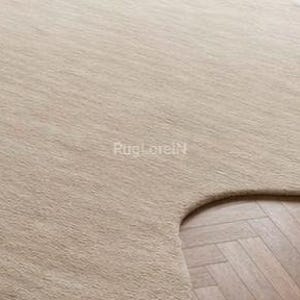 May include: A beige, uniquely shaped area rug with a soft, textured surface. The rug's irregular edges contrast with the herringbone pattern of the wooden floor it partially covers. The rug is a neutral color.