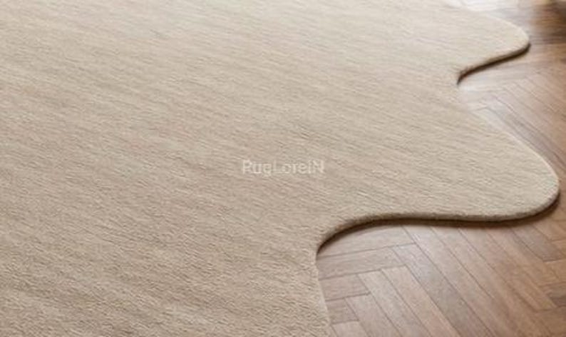 May include: A beige, uniquely shaped area rug with a soft, textured surface. The rug's irregular edges contrast with the herringbone pattern of the wooden floor it partially covers. The rug is a neutral color.
