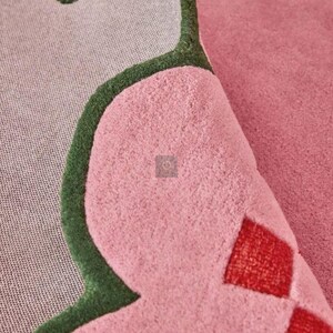 May include: Close-up of a pink rug with a unique shape and a green border. The rug has a soft, textured surface and a gray backing. Red diamond shapes are visible on the rug's surface.