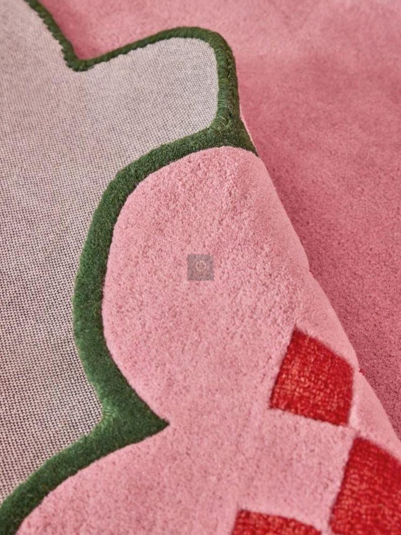 May include: Close-up of a pink rug with a unique shape and a green border. The rug has a soft, textured surface and a gray backing. Red diamond shapes are visible on the rug's surface.