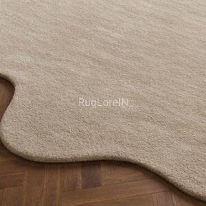 May include: A beige, organically shaped rug with a soft, textured surface. The rug's unique form contrasts with the dark brown wooden floor beneath it. The text "RugLoreIN" is visible on the rug's surface.