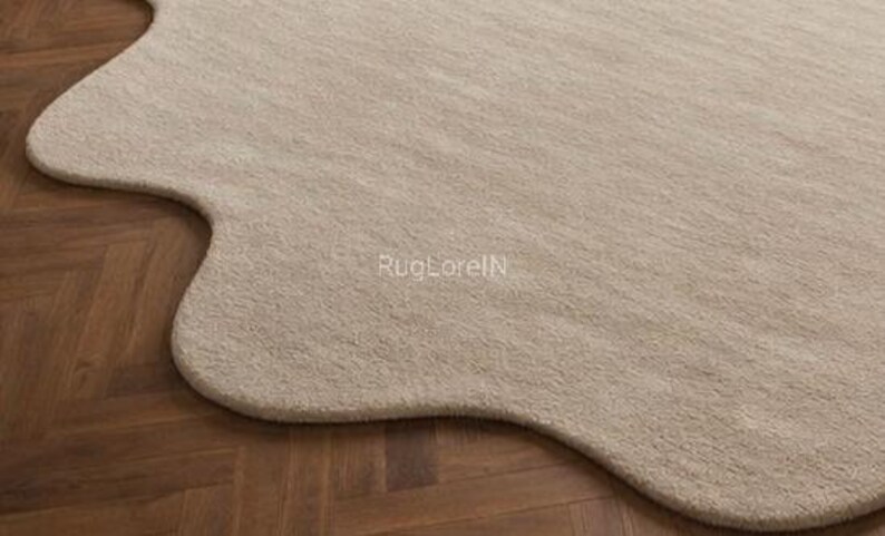 May include: A beige, organically shaped rug with a soft, textured surface. The rug's unique form contrasts with the dark brown wooden floor beneath it. The text "RugLoreIN" is visible on the rug's surface.