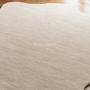 May include: A large, neutral-toned area rug with an organic, irregular shape. The rug has a soft, textured appearance, with subtle variations in color. The surface appears to be made of a woven material. The rug is placed on a hardwood floor.