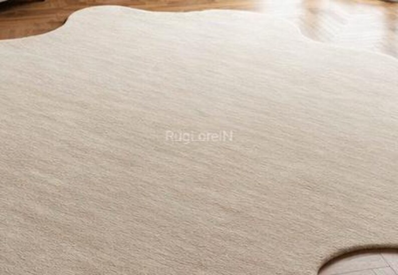 May include: A large, neutral-toned area rug with an organic, irregular shape. The rug has a soft, textured appearance, with subtle variations in color. The surface appears to be made of a woven material. The rug is placed on a hardwood floor.
