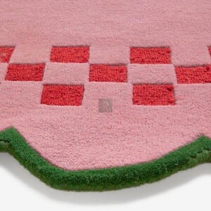 May include: A pink rug with a checkerboard pattern in red and pink. The rug has a wavy edge trimmed with green. The rug is made of a soft, plush material.