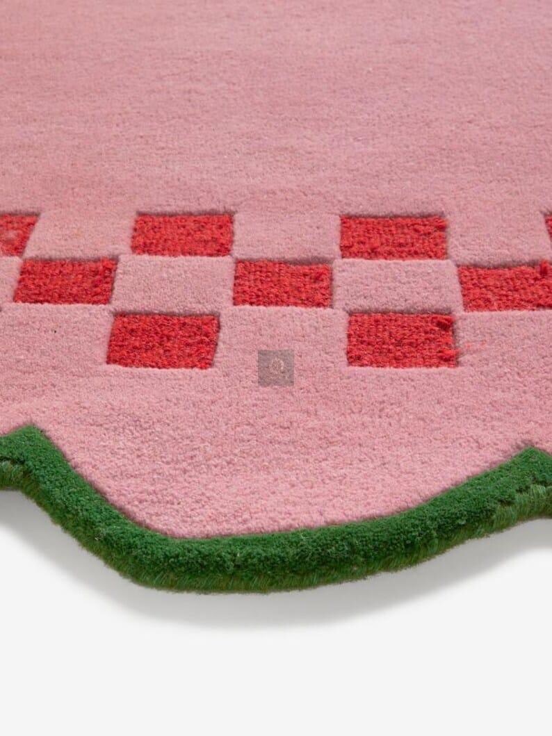 May include: A pink rug with a checkerboard pattern in red and pink. The rug has a wavy edge trimmed with green. The rug is made of a soft, plush material.