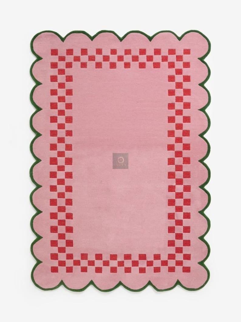 May include: A rectangular pink rug with a scalloped green border. The rug features a red checkered pattern around the edges and a solid pink center. The rug is designed for indoor use.