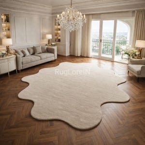 May include: A large, beige, irregularly shaped rug dominates a room with hardwood flooring. The rug's unique design adds a soft contrast to the room's elegant decor, which includes a sofa, chairs, and a crystal chandelier. The text "RugLoreIN" is visible on the rug.