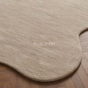 May include: A beige, irregularly shaped rug with a soft, textured surface. The rug's edge curves gently, contrasting with the wooden floor's straight lines. The text "RugLoreIN" is visible on the rug's surface.