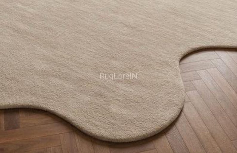 May include: A beige, irregularly shaped rug with a soft, textured surface. The rug's edge curves gently, contrasting with the wooden floor's straight lines. The text "RugLoreIN" is visible on the rug's surface.