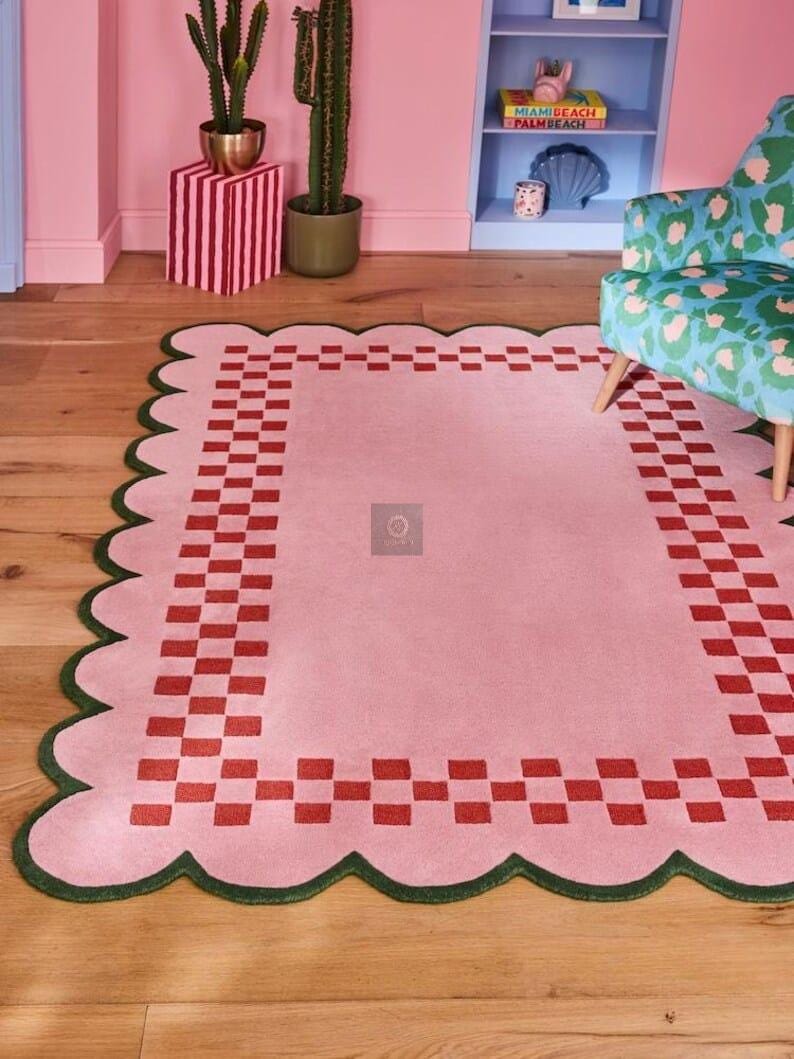 May include: A pink rectangular rug with a scalloped green border and a red checkerboard pattern. The rug is placed on a wooden floor, with a chair and a bookshelf in the background. The rug's dimensions are not visible.