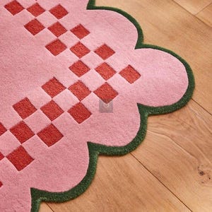 May include: A pink rug with a scalloped edge and a green border. The rug features a repeating pattern of red squares. The rug is made of a soft material and is designed for home decor.