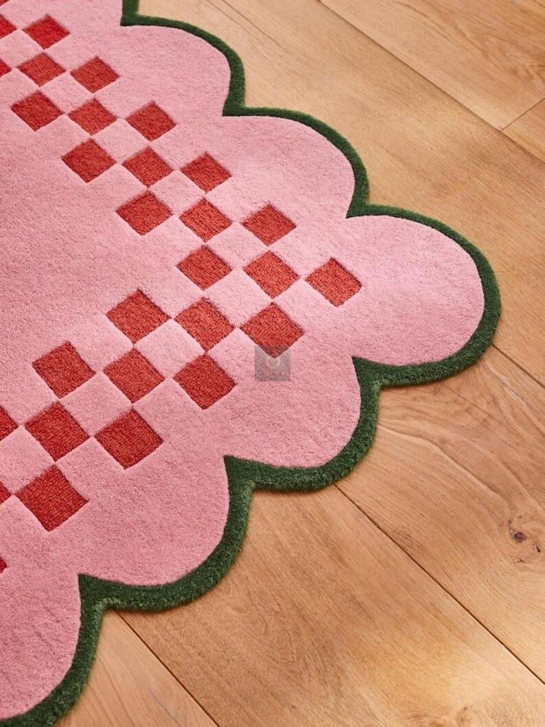 May include: A pink rug with a scalloped edge and a green border. The rug features a repeating pattern of red squares. The rug is made of a soft material and is designed for home decor.