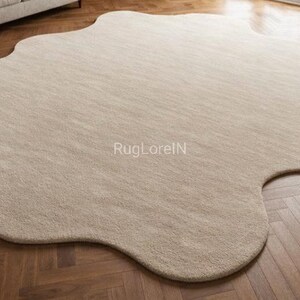 May include: A large, beige, organically shaped area rug. The rug has a soft, textured appearance and is placed on a dark wood floor. The rug's unique shape adds a modern touch to the room's decor. The text "RugLorelN" is visible on the rug.