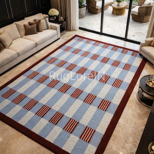May include: A rectangular rug featuring a blue, white, and burgundy chequered design. The rug has a burgundy border and is placed on a marble floor in a living room. The rug's pattern provides a geometric element to the room's decor.