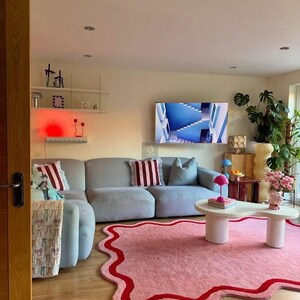 May include: A modern living room with a light gray sectional sofa, a pink rug with a red border, and a white coffee table. Decorative pillows with red and white stripes add color. A TV displays a blue image.