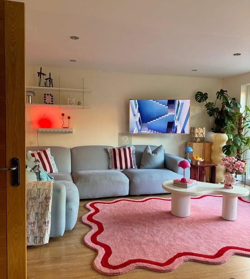 May include: A modern living room with a light gray sectional sofa, a pink rug with a red border, and a white coffee table. Decorative pillows with red and white stripes add color. A TV displays a blue image.
