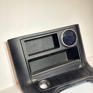 Könnte beinhalten: Schwarzes Kfz-Armaturenbrett-Bauteil mit einem Messgerät und Ablagefächern. Das Messgerät hat ein schwarzes Zifferblatt mit roten Markierungen und einem Zeiger. Die Komponente hat eine strukturierte Oberfläche und eine kreisförmige Öffnung.