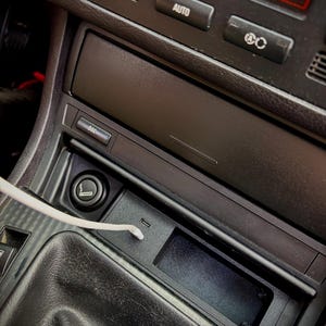 Op de afbeelding: Auto-interieurconsole met een sigarettenaansteker, een klein opbergvak en een USB-poort. De temperatuurweergave toont 20,0°C. De console is zwart met zilveren accenten.