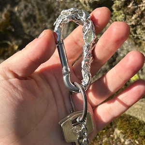 Può includere: Un portachiavi color argento con un moschettone decorativo e strutturato. Il moschettone è attaccato a una catena che tiene diverse chiavi. Il design del moschettone ha una forma organica e fluida con un accento di pietra preziosa.