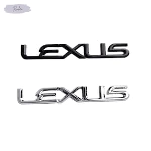 Puede incluir: Dos emblemas de coche Lexus. El emblema superior es negro y el inferior es cromado. Ambos emblemas tienen la palabra "LEXUS" en una fuente estilizada. El fondo es blanco.