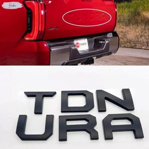 Puede incluir: Un conjunto de letras adhesivas individuales en negro mate que forman la palabra TUNDRA, diseñadas para la puerta trasera de un vehículo. Las letras presentan una fuente de estilo bloque con bordes definidos, dispuestas sobre una lámina blanca.