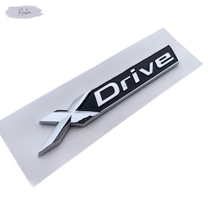 Peut inclure: Un emblème "Drive" chromé et noir avec un design "X" stylisé. Le mot "Drive" est en blanc, sur fond noir. L'emblème est sur un support rectangulaire blanc.