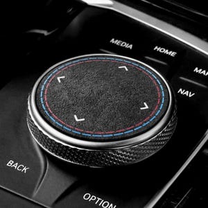 以下が含まれることがあります： 車のコントロールノブのクローズアップ。黒いノブは、テクスチャ加工された表面とシルバーの金属リムを備えています。青と赤の円形アクセントと白い矢印が付いています。「MEDIA」、「HOME」、「NAV」、「BACK」、「OPTION」と書かれたボタンがあります。