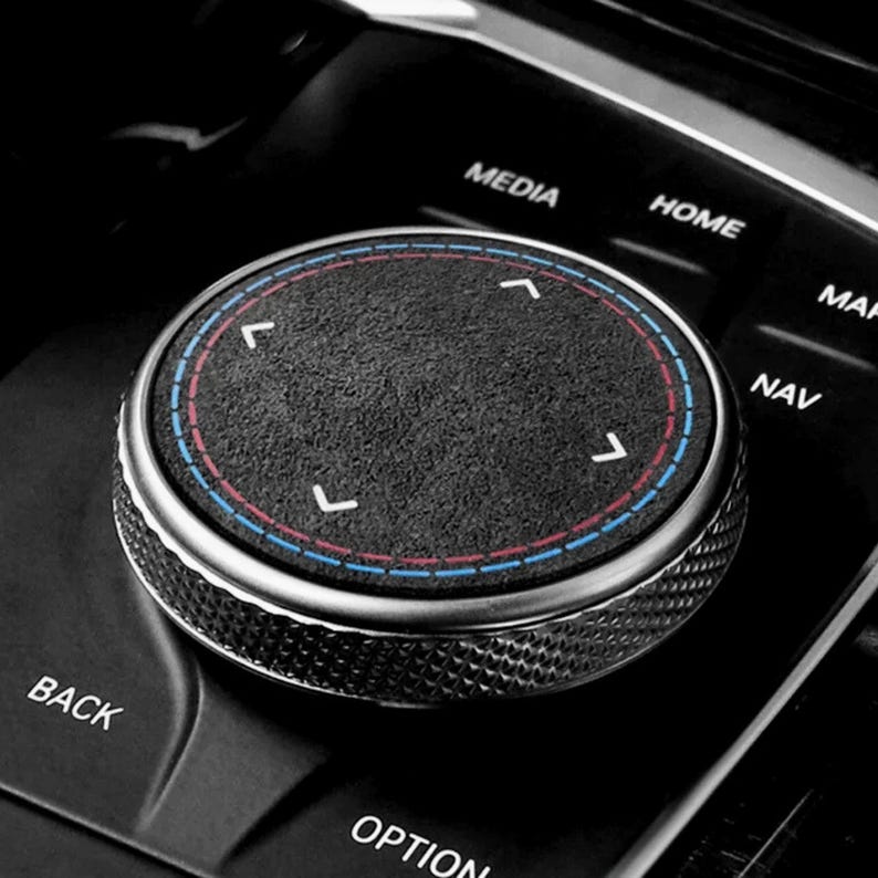 May include: Close-up of a car's control knob. The knob is black with a textured surface and a silver metal rim. It features a blue and red circular accent and white directional arrows. The surrounding buttons are labeled "MEDIA", "HOME", "NAV", "BACK", and "OPTION".