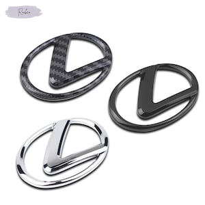 Puede incluir: Tres insignias de emblema de coche. Una tiene un patrón de fibra de carbono, otra es de color negro brillante y la otra es cromada. Cada emblema presenta una letra "L" estilizada dentro de un marco ovalado. Las insignias se muestran sobre un fondo blanco.
