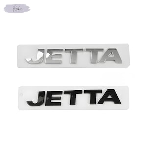 May include: Two car emblems with the word "JETTA" in chrome and black. Each emblem is mounted on a white rectangular base. The chrome emblem is at the top, and the black emblem is below it. The background is white.