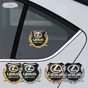 Op de afbeelding: Een verzameling Lexus Motors auto-emblemen in goud, zilver en zwart. Elk embleem toont het Lexus-logo en de woorden "Lexus Motors" in een schildontwerp, omringd door een lauwerkrans. De emblemen worden getoond op een autoruit.