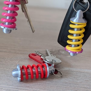 Könnte beinhalten: Schlüsselanhänger mit einem Miniatur-Stoßdämpfer-Design. Die Schlüsselanhänger haben eine silberfarbene Ober- und Unterseite mit einer leuchtend farbigen Feder in Pink, Gelb und Rot. Die Schlüsselanhänger sind an Schlüsseln und einem Autoschlüsselanhänger befestigt.
