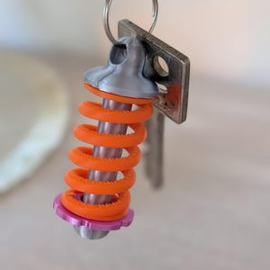 Puede incluir: Un llavero impreso en 3D con una parte superior plateada, un resorte naranja vibrante y una base rosa. El llavero está unido a un anillo de metal con una llave y una etiqueta de metal. El diseño es una versión lúdica de un componente mecánico.