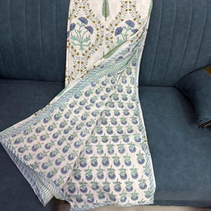 Könnte beinhalten: Ein weißes Textil mit einem Blumenmuster in Blau, Grün und Gold ist über ein blaues Sofa drapiert. Das Textil zeigt ein sich wiederholendes Muster aus blauen Blumen und grünen Blättern mit einem gestreiften Rand.